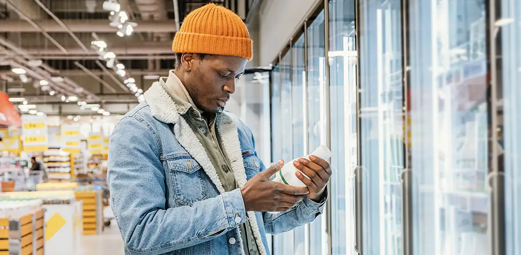 A man wearing an orange beanie and denim jacket reads the label on a product while shopping in the refrigerated section of a grocery store.