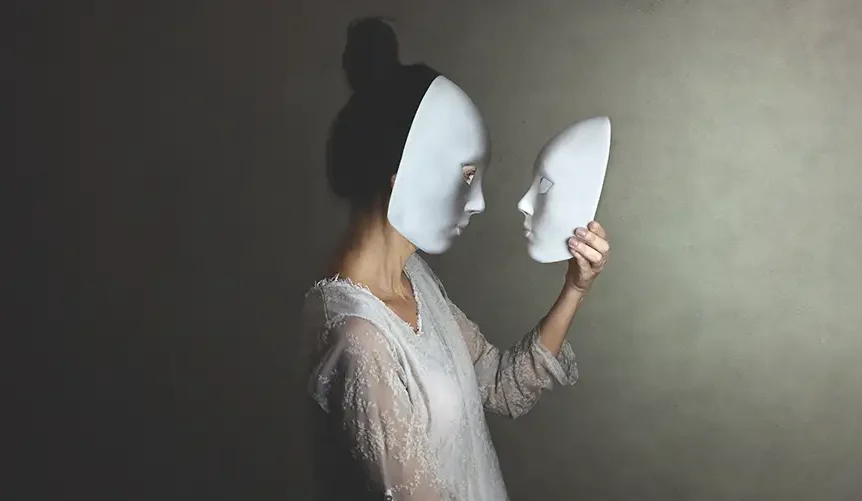 Eine Frau steht im Profil. Sie trägt eine weiße Maske, hält eine gleiche weiße Maske in den Händen und schaut darauf. Sie trägt ein weißes, geschnürtes Kleid.