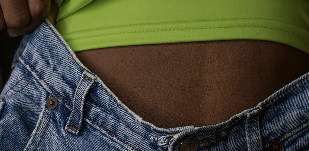 Persoană purtând blugi din denim albastru, cu talia îndepărtată de stomac, cu un top verde vizibil.