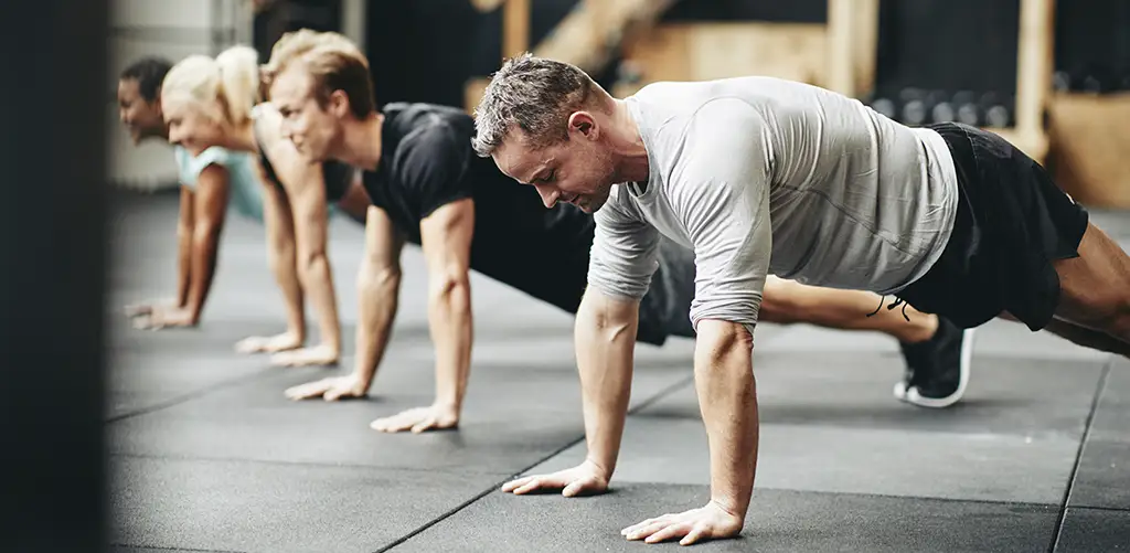 Un groupe de personnes fait de l'exercice ensemble dans une salle de sport, en faisant des pompes sur le sol. L'accent est mis sur un homme en chemise d'entraînement grise à l'avant, avec d'autres participants alignés derrière lui.
