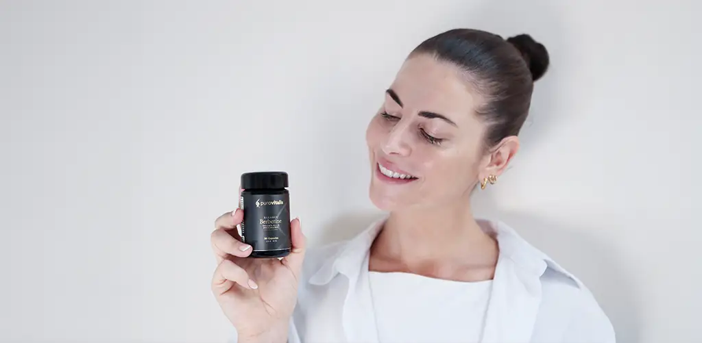 Una donna sorridente con i capelli scuri raccolti in uno chignon, che indossa una camicia bianca, tiene in mano una bottiglia black di Purovitalis Berberine Balance su uno sfondo chiaro.