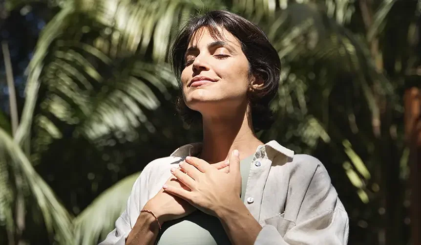Una mujer está de pie al aire libre, con los ojos cerrados y una sonrisa serena, con las manos sobre el pecho mientras respira profundamente. La luz del sol se filtra a través de la vegetación tropical que la rodea, creando una atmósfera tranquila y plena.