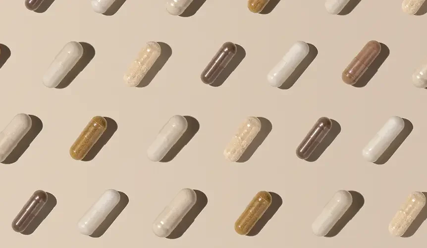 Cápsulas de suplementos dispuestas en un ordenado patrón repetitivo sobre un fondo beige, en diversos tonos de blanco, tostado y marrón, que proyectan sombras suaves.