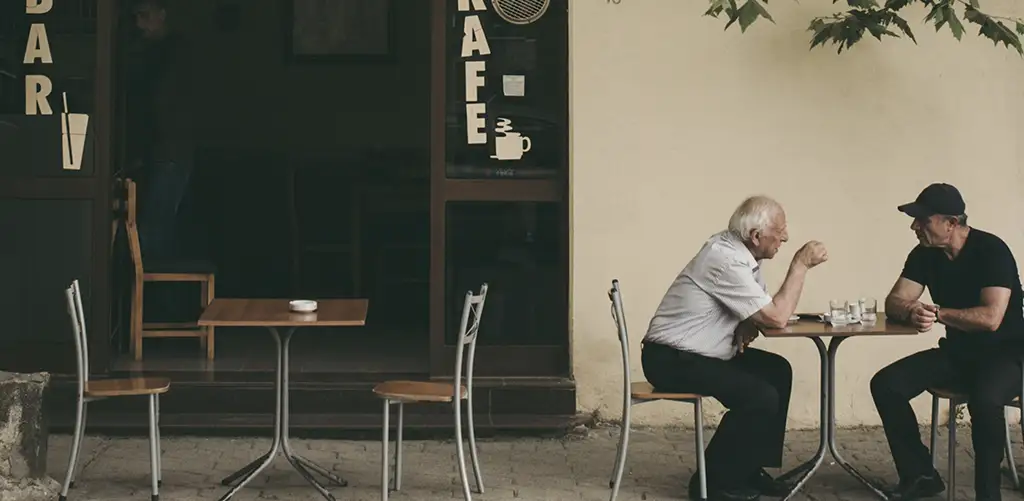 Twee oudere mannen zitten aan een klein tafeltje op een terrasje en voeren een gesprek, wat de sociale verbondenheid en kameraadschap op latere leeftijd illustreert.