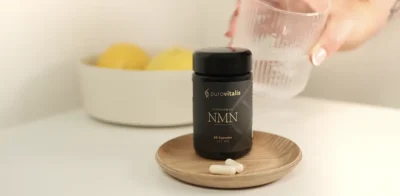 Purovitalis Liposomal NMN kosttilskudsflaske på en lille træplade med kapsler foran, placeret på et hvidt bord, med en hånd, der holder et glas vand i baggrunden.