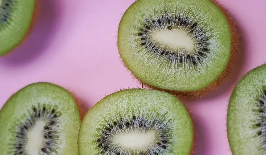 Primer plano de varios kiwis verdes cortados en rodajas y dispuestos sobre un fondo rosa, en el que se aprecia su pulpa de color verde brillante, sus centros blancos y sus pequeñas semillas negras dispuestas en forma radial.