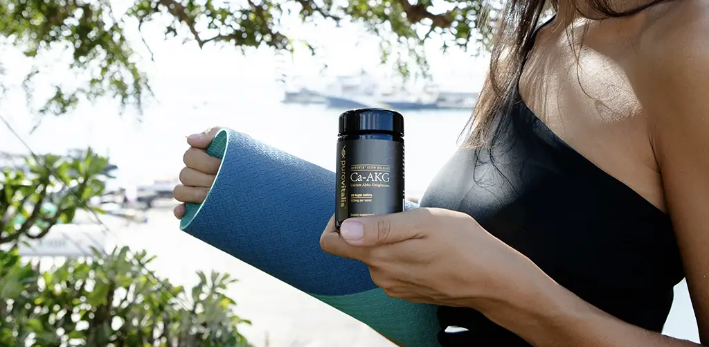Frau hält im Freien eine Flasche mit Calcium-Alpha-Ketoglutarat (Ca-AKG)-Nahrungsergänzungsmittel auf einer Yogamatte.