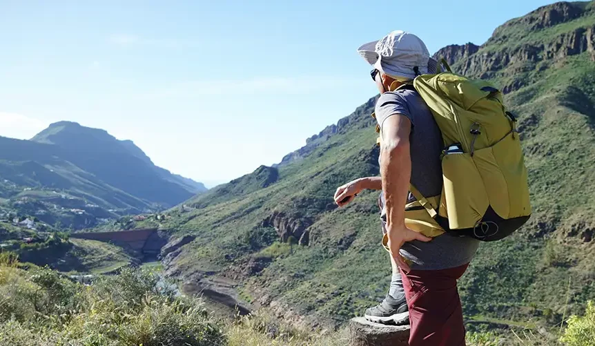 Man hiking in a mountainous landscape with a large backpack, looking out over a sunny valley during an outdoor trekking adventure.