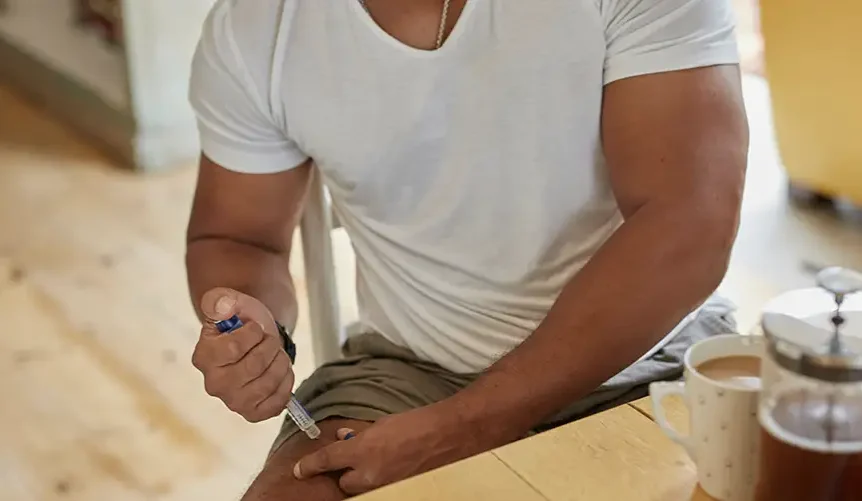 A muscular man administering a GLP-1 injection pen into his thigh while seated at a wooden table next to a coffee cup.