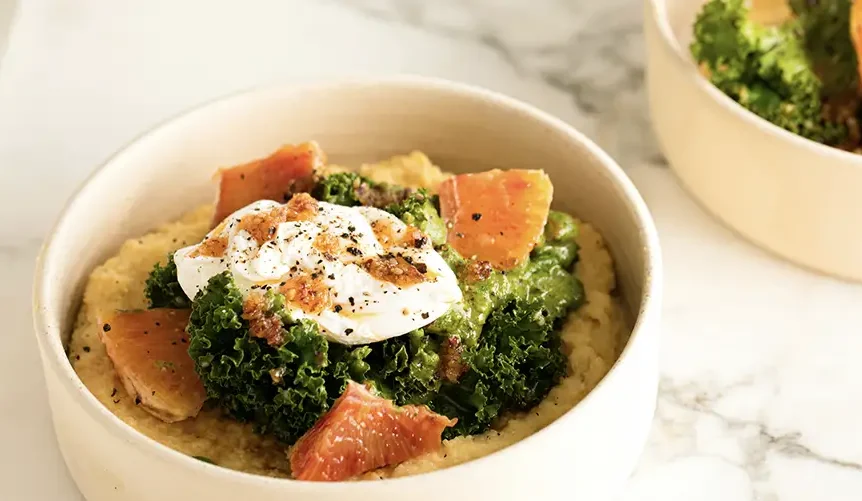 A healthy bowl of polenta topped with sauteed kale, blood orange slices, and a poached egg on a marble surface.